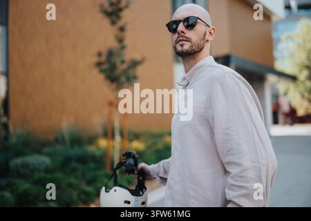 Homme chauve portant des lunettes de soleil tient un casque tout en marchant à côté de son scooter électrique. Portrait urbain décontracté d'un voyageur confiant dans une ville moderne s Banque D'Images