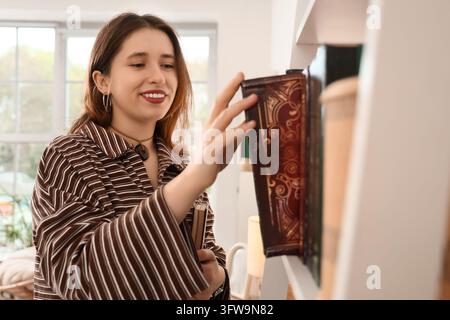 Éducation et apprentissage tout au long de la vie, jeune femme choisissant un livre sur une étagère de bibliothèque à domicile pour la lecture et le développement personnel dans un salon lumineux Banque D'Images