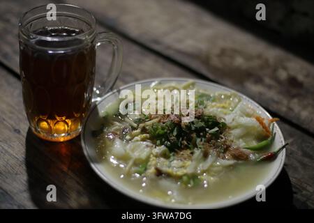 Soupe de nouilles traditionnelle indonésienne Bakmi Jawa servie avec des légumes, des herbes et du poulet émincé, accompagnée d'un verre de thé chaud sur du bois rustique Banque D'Images