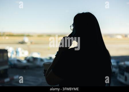 Femme en silhouette de profil, téléphonant sur un téléphone portable près de la fenêtre d'un terminal d'aéroport, regardant les avions sur la piste Banque D'Images