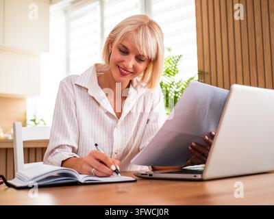 Travailleuse à distance concentrée écrivant dans un carnet tout en vérifiant des documents à son bureau à domicile. Banque D'Images