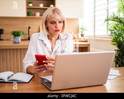 Femme inquiète vérifiant une notification importante tout en travaillant à domicile avec un ordinateur portable et des papiers. Banque D'Images