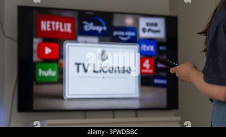 Une personne pointe une télécommande vers une télé intelligente affichant des icônes d'applications de streaming et un message de licence TV, illustrant les abonnements de divertissement à domicile et la télévision Banque D'Images
