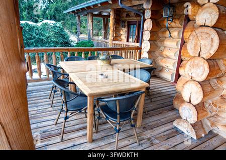 Terrasse couverte et confortable d'une cabane rustique en rondins avec tables en bois, chaises en osier et architecture en bois naturel entourée d'arbres à feuilles persistantes Banque D'Images