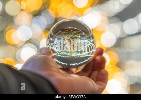 Vue d'un sapin de Noël reflété dans une boule de cristal tenue dans une main devant un fond de lumières bokeh festives Banque D'Images