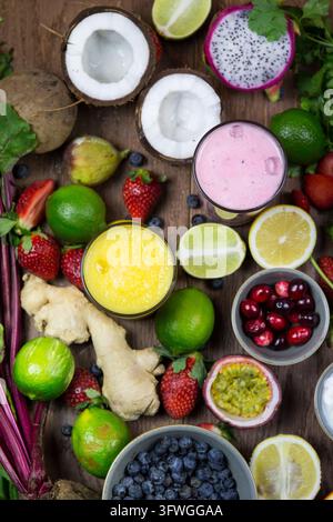 Disposition en plan vertical de fruits tropicaux, baies et gingembre entourant des smoothies roses et jaunes sur une surface en bois rustique. Banque D'Images