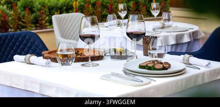 Une table de dîner en plein air élégamment dressée présente des nappes blanches, des verres à vin remplis de vin rouge et des assiettes de hors-d'œuvre gastronomiques. Banque D'Images