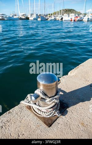 Un taquet d'amarrage en acier inoxydable brillant fixé avec une corde blanche épaisse sur un quai en béton ensoleillé, avec une eau de mer bleu profond et des bateaux en arrière-plan. Banque D'Images