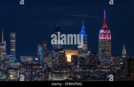Vue aérienne de la skyline de New York City la nuit avec l'Empire State Building illuminé en rouge et bleu et une pleine lune se levant derrière les gratte-ciel Banque D'Images