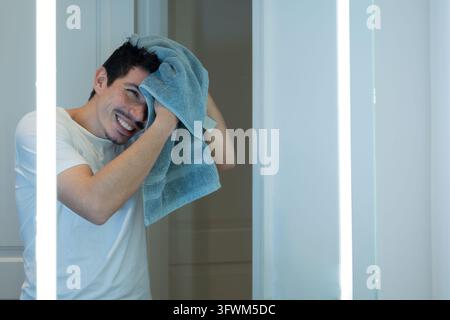 Homme joyeux séchant ses cheveux avec une serviette devant un miroir lumineux après avoir pris un bain Banque D'Images