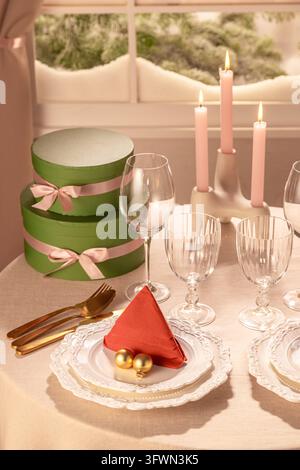 Gros plan en hauteur d’un couvert de Noël mettant en valeur une serviette rouge en forme d’arbre, des boules dorées, des verres à vin en cristal, des bougies roses allumées et des ronds empilés Banque D'Images