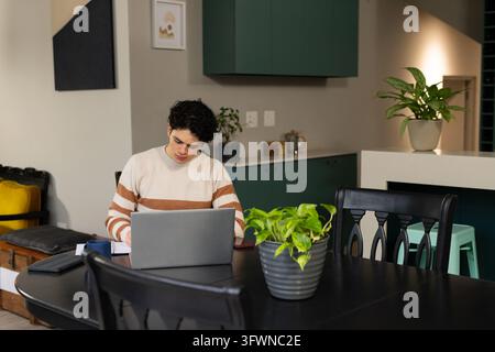 Homme afro-américain début de la vingtaine travaillant sur un ordinateur portable à la table à manger avec un cahier, un stylo, un pull crème. Bureau à domicile, ordinateur, plantes, chaleur, éclairage doux Banque D'Images