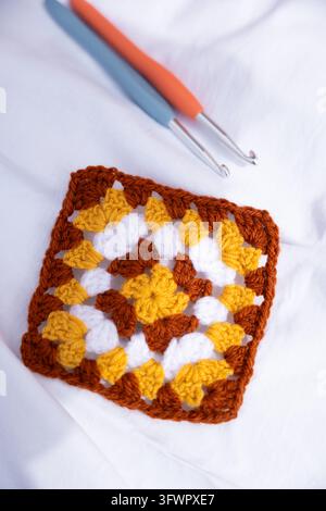 Carré granny fait à la main au crochet avec de la laine brune, jaune et blanche, présenté sur un tissu doux avec deux crochets, mettant en valeur un motif floral texturé an Banque D'Images