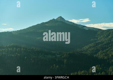 Pic acéré de calcaire s'élevant au-dessus d'une forêt dense à feuillage persistant avec un petit chalet dans une clairière ensoleillée sous un ciel bleu clair et une lumière douce du matin qui projette des ombres Banque D'Images