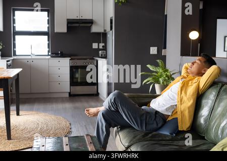 Homme asiatique portant une veste moutarde, allongé sur un canapé vert avec un coussin à la maison, espace de copie Banque D'Images
