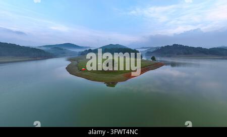 Vue aérienne d'un arbre solitaire et d'une petite tente sur une péninsule herbeuse entourée par les eaux calmes du lac Tuyen Lam à Da Lat, Lam Dong, Vietnam. Banque D'Images