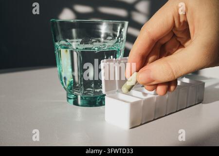 Main prenant une capsule verte dans une boîte à pilules à côté d'un verre d'eau Banque D'Images