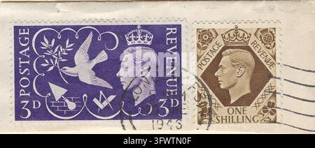 Histoire postale de la Grande-Bretagne de 1946 sur un fragment d’enveloppe d’origine, affranchie d’un timbre commémoratif violet de 3d Paix et Victoire et d’un timbre définitif marron d’un shilling du roi George VI, tous deux portant un cachet de 1946. Le motif de 3d représente une colombe portant une branche d’olivier à côté du monarque couronné, avec des symboles de reconstruction après la Seconde Guerre mondiale. L’émission adjacente d’un shilling de 1939 porte l’inscription POSTAGE REVENUE. Les perforations d’origine et les marquages postaux ronds noirs et en forme d’onde préservent un enregistrement distinctif de la paix et de la communication d’après-guerre britanniques. Banque D'Images