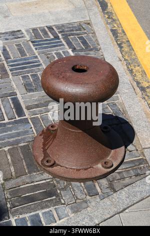 Une bittement d'amarrage en fonte recouvert de rouille sur le pavé en brique extérieur, avec un trou rond sur le dessus et des trous de boulon à la base, montrant un style industriel rétro Banque D'Images