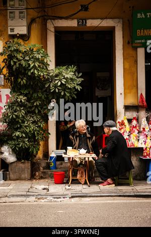 Hanoï, Vietnam - 16 janvier 2026 : Deux hommes âgés sont assis et discutent à une petite table devant l’entrée d’un bâtiment jaune avec des plantes en pot et des couleurs vives… Banque D'Images