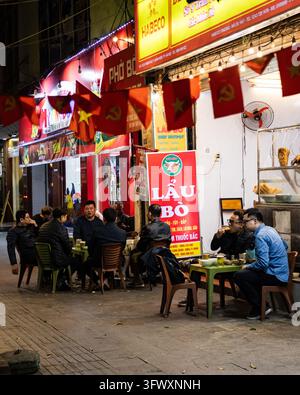 Hanoï, Vietnam - 31 décembre 2025 : Groupe d'hommes assis et mangeant à de petites tables devant un restaurant de rue de nuit avec du rouge et du jaune… Banque D'Images