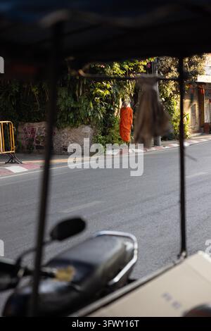 Chiang Mai, Thaïlande - 17 janvier 2026 : Un moine bouddhiste en robes orange marche le long d'une rue bordée de verdure, vu à travers le cadre d'un… Banque D'Images