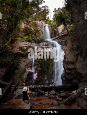 Pai, Thaïlande - 19 janvier 2026 : Une personne portant un chapeau est assise sur des rochers et prend une photo d'une cascade entourée d'une végétation luxuriante et... Banque D'Images