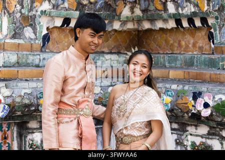 Bangkok, Thaïlande - 1er février 2026 : Un homme et une femme thaïlandais souriants, vêtus de tenues traditionnelles roses coordonnées, se tiennent devant un lieu coloré… Banque D'Images