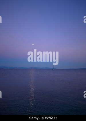 Un voilier solitaire repose sur une mer violette et calme sous une lune brillante avec un reflet de lune léger et des montagnes lointaines en Grèce. Banque D'Images