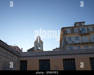 Un clocher blanc et jaune avec deux cloches s'élève au-dessus d'un toit de tuiles et d'un vieux bâtiment aux volets fermés dans la ville de Corfou, en Grèce. Banque D'Images
