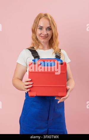 Femme en salopette bleue souriant et tenant une boîte à outils rouge à deux mains, présentant du matériel pour les réparations à domicile, la construction ou les projets de bricolage Banque D'Images