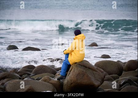 Femme assise sur un gros rocher observant les puissantes vagues de la mer de Norvège à la plage d'Unstad, Lofoten, Norvège, entourée par une côte rocheuse hivernale par un temps côtier arctique froid Banque D'Images
