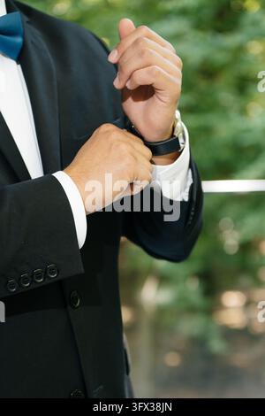 Gros plan de mains soignées ajustant la manchette d'une chemise avec une montre-bracelet, un costume noir et un nœud papillon bleu Banque D'Images