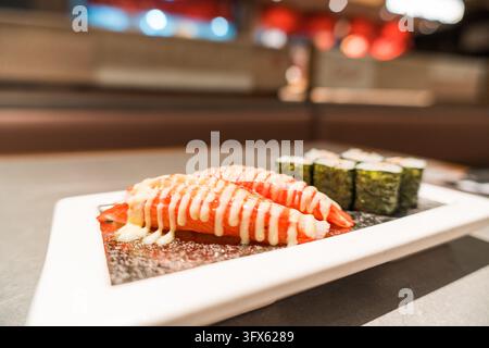 Plateau de sushi japonais fraîchement préparé comprenant deux pièces de nigiri au saumon recouvertes d'une sauce crémeuse et une rangée de roulés maki enveloppés dans du nori vert, servis sur une assiette en céramique rectangulaire blanche garnie de graines de sésame et de wasabi, dans un intérieur de restaurant haut de gamme moderne avec un éclairage ambiant chaleureux et un bokeh flou d'arrière-plan créé par des lumières décoratives rouges et bleues Banque D'Images