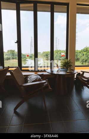 Intérieur de café minimaliste moderne avec de grandes baies vitrées surplombant un plan d'eau tranquille et de la verdure, comprenant des chaises en rotin tissé, une table ronde en bois avec un ordinateur portable ouvert et une plante en pot, un sol carrelé en céramique sombre et une lumière naturelle du jour illuminant l'espace dans une atmosphère sereine et productive Banque D'Images