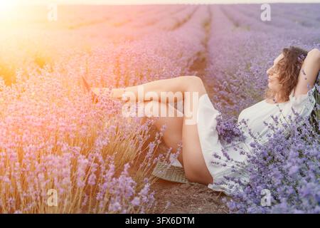 Femme se relaxant dans un champ de lavande en été, scène de nature avec une belle dame en robe blanche reposant paisiblement parmi des fleurs violettes en fleurs au coucher du soleil Banque D'Images