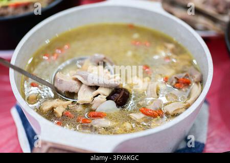 Soupe de poulet aux herbes chinoises traditionnelle brûlante avec baies de goji, champignons séchés, jujube et abats de porc, servie dans un pot en céramique blanche avec une cuillère en métal, mijotant sur une nappe rouge dans un cadre de repas maison Banque D'Images