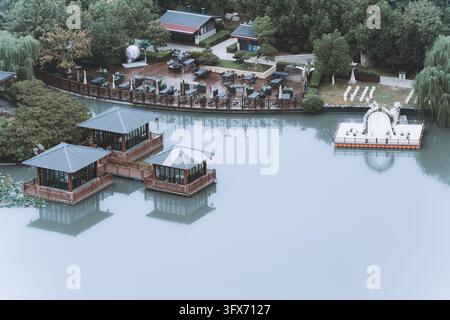Vue aérienne de pavillons flottants de style chinois traditionnel sur un lac calme avec une décoration de mariage au bord du lac comprenant une arche florale blanche et des rangées de chaises, entourée d'une végétation luxuriante incluant des saules pleureurs et des jardins bien entretenus dans un cadre de parc serein, probablement à Hangzhou, en Chine Banque D'Images