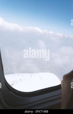 Vue aérienne depuis une fenêtre d'avion montrant l'aile blanche de l'avion s'étendant dans un ciel bleu éclatant au-dessus d'une couche de nuages cumulus dispersés, avec un paysage urbain à peine visible en dessous à travers des conditions atmosphériques brumeuses Banque D'Images