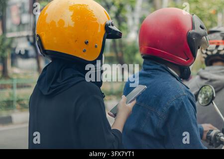 Vue rapprochée de dos de deux motards portant des casques jaunes et rouges sur une route, un passager tenant un smartphone Banque D'Images