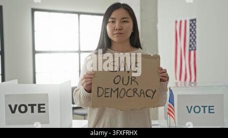 Femme tenant un panneau en carton lisant «sauvez notre démocratie» devant des isoloirs et une urne dans une salle de vote à l'intérieur d'un bâtiment ; détermination civique Banque D'Images