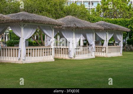 Range de cabanes de luxe au toit de chaume avec rideaux blancs sur une pelouse verte dans un resort Banque D'Images