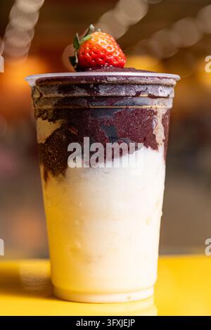 Prise en studio d'un bol d'acai dans une tasse en plastique garni de fraise. La texture crémeuse contraste avec l'acai sombre sur un fond chaud. Représente un ea sain Banque D'Images