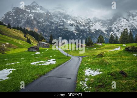Route étroite et sinueuse sur le pâturage alpin avec de jolies cabanes en bois et des montagnes brumeuses spectaculaires en arrière-plan, Murren, vallée de Lauterbrunnen, Suisse Banque D'Images