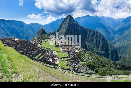 Les terrasses, places et structures en pierre emblématiques de Machu Picchu sont entourées de pics montagneux verts et acérés sous un ciel bleu. Banque D'Images