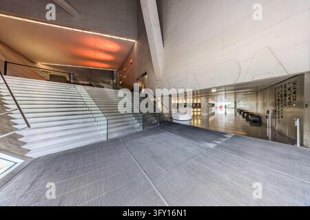 La Casa da Música à Porto, Portugal, possède un large escalier avec des marches pâles et une balustrade en verre dans son hall. Des murs en béton avec des surfaces inclinées entourent l'espace, et le plafond au-dessus des escaliers brille en orange. De grandes lettres sur le mur indiquent « CASA DA ». Des panneaux sur un mur métallique pointent vers « RESTAURANTE », « SALA 1 » et « CAFÉ », entre autres (En anglais : Restaurant, Salle 1, Café). Rem Koolhaas et le bureau OMA ont conçu la salle de concert. Elle a ouvert ses portes en avril 2005. La ville a été Capitale européenne de la culture en 2001, et le bâtiment a été créé pour cette année-là. Banque D'Images