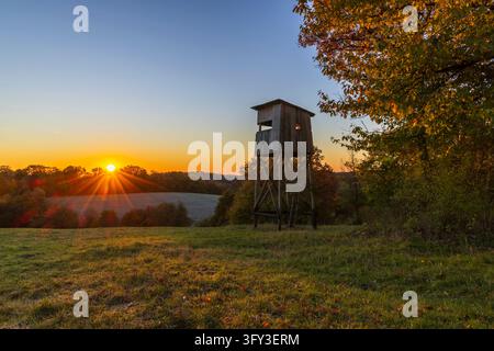 Stand de chasse en bois surélevé au-dessus d'un champ, avec le soleil d'automne se couchant sur des collines boisées Banque D'Images