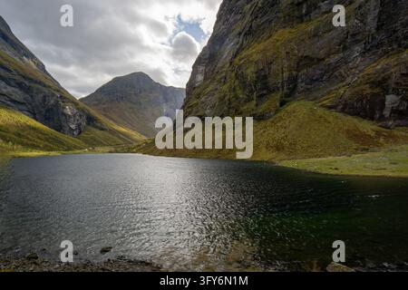 Croisière à travers le Hjørundfjorden, en Norvège, entourée de falaises montagneuses abruptes, de vallées verdoyantes, de cascades et d'une eau sombre et réfléchissante sous un ciel nuageux. Banque D'Images