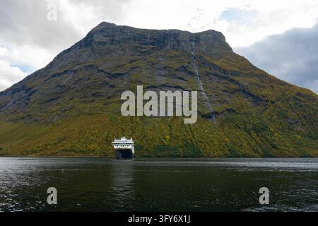 Bateau de croisière naviguant sous une montagne abrupte striée de cascades dans le fjord de Hjørund, en Norvège, entouré par une forêt d'automne et des eaux sombres du fjord. Banque D'Images