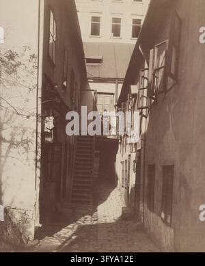 9e arrondissement, Badgasse 3 - Zu den 3 weißen Tauben - Vue sur la cour, August Stauda (1861–1928), Photographe, photographies, vers 1905, passe-partout 42×32 cm, taille de l'image 27,7×21,8 cm, Architecture, Sites et structures disparus, 9e arrondissement : Alsergrund, enfant, escalier, façade de la cour intérieure, avec des personnes, Vienne Autriche Collection Kabinett, Une ruelle pavée étroite entre deux bâtiments montre une personne debout sur un balcon extérieur au sommet d'une courte volée d'escaliers dans un milieu urbain Banque D'Images